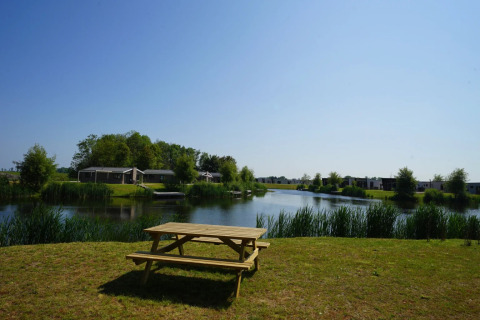 Picnic table by a lake with glamping lodges in the background at a holiday park under a clear blue sky.