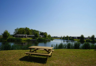 Picnic table by a lake with glamping lodges in the background at a holiday park under a clear blue sky.