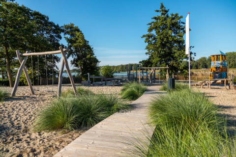 Aire de jeux sur le sable dans un parc de vacances au bord d’un lac, avec balançoires et structures en bois.