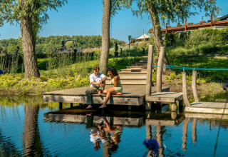 To personer sidder på en træbro ved en sø i et feriepark med glamping, omgivet af frodig natur.