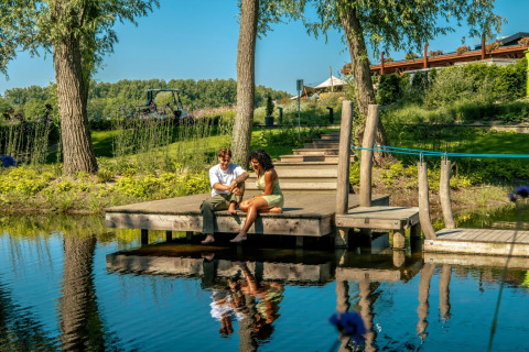 Twee mensen zitten op een houten steiger bij een vijver in een vakantiepark met glamping, omgeven door groen.