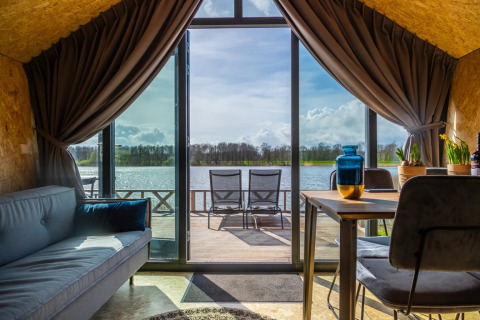 Vista desde una cabaña glamping en un parque vacacional con sofá, mesa y terraza frente a un lago soleado.