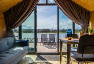 Vue d’une cabane glamping dans un parc de vacances, avec canapé, table et terrasse surplombant le lac ensoleillé.