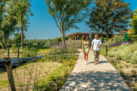 Pareja caminando de la mano por un sendero de madera en un parque vacacional de glamping con jardines verdes.
