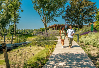 Stel loopt hand in hand over een houten pad in een vakantiepark met glamping, omgeven door groene tuinen.