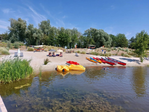 Playa con kayaks, botes de pedales, hamacas y huéspedes relajándose, rodeada de árboles en parque glamping.