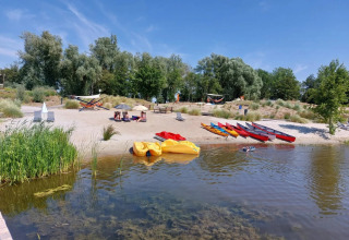 Strand med kajakker, pedalbåde, hængekøjer og gæster, omgivet af træer på feriepark med glamping.