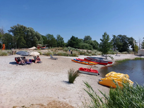 Holiday park beach with glamping, people lounging under umbrellas, kayaks and pedal boats by the water.