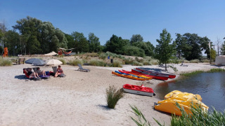 Playa de parque vacacional con glamping, gente bajo sombrillas, kayaks y botes a pedales junto al agua.