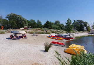 Vakantieparkstrand met glamping, mensen onder parasols, kajaks en waterfietsen naast het water.