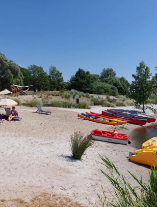 Plage d’un parc de vacances avec glamping, personnes sous parasols, kayaks et pédalos près de l’eau.