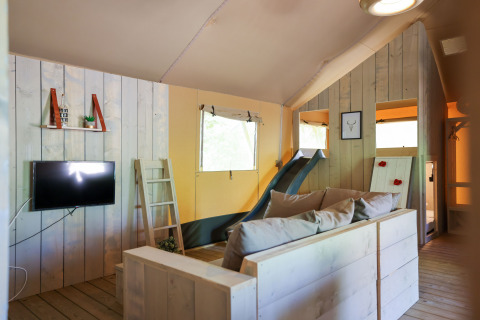 Vista interna di una tenda glamping con pannelli in legno, divano, TV, scala e scivolo.