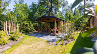 Charming Heide Lodge tiny house with porch set in a lush, green garden surrounded by tall trees.