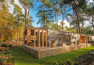 Cabane de glamping moderne avec grande terrasse dans un parc de vacances entouré de hauts arbres.