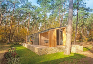 Cabane de glamping moderne, bois et pierre, nichée dans une forêt ensoleillée d’un parc de vacances.