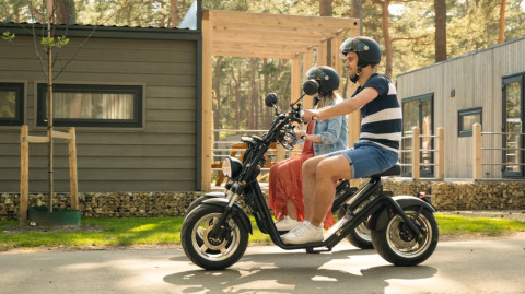 Pareja montando en scooter eléctrico frente a cabañas en un parque de vacaciones con glamping y naturaleza.