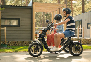 Couple à scooter électrique devant des cabanes dans un parc de vacances avec glamping, nature environnante.