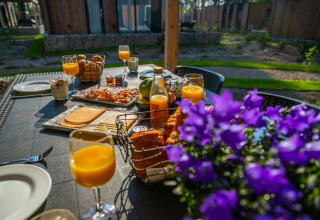 Ontbijt in de buitenlucht met fruitsap, kaas, brood en bloemen bij een glamping vakantiepark.