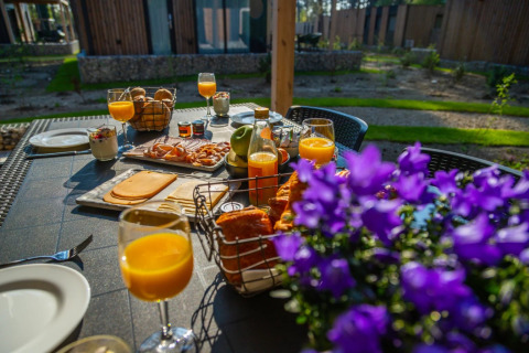 Ontbijt buiten met sap, kaas, brood en bloemen op tafel bij een glamping vakantiepark in de natuur.