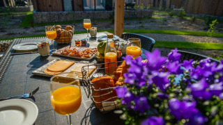 Frühstück im Freien mit Saft, Käse, Brot und Blumen auf dem Tisch im Glamping-Urlaubspark in Naturumgebung.