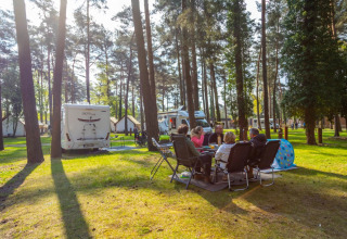 Parco vacanze con glamping, camper e persone che si rilassano insieme sotto alberi alti durante il giorno.