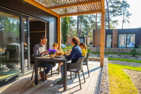 Familia desayunando en la terraza de un glamping con cabañas modernas y naturaleza al fondo, día soleado.