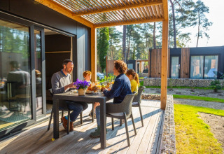 Gezin geniet van ontbijt op het terras bij een glampingpark, met moderne chalets en groene natuur.
