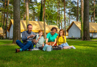 Famille pique-niquant sur l'herbe devant des tentes glamping dans un parc de vacances entouré de forêt.