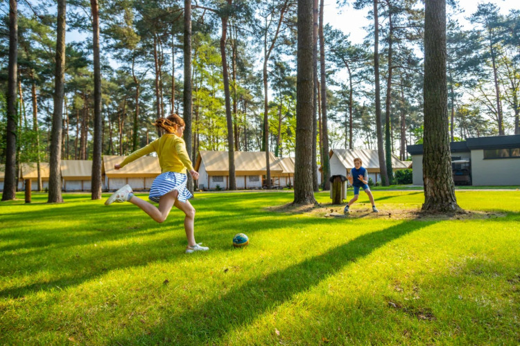 Two children play soccer on a grassy field surrounded by trees and glamping tents at a holiday park.