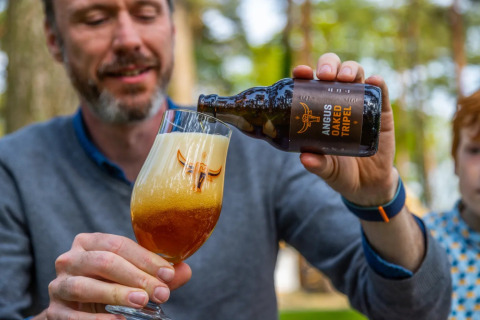 Homme versant une bière Angus Oaked Tripel dans un verre dans un parc de glamping, forêt derrière.