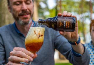 Homme versant une bière Angus Oaked Tripel dans un verre dans un parc de glamping, forêt derrière.