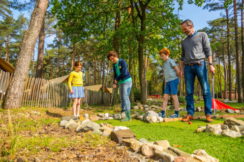Una familia juega al minigolf al aire libre en un parque vacacional con tiendas glamping y bosque detrás.