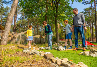 Une famille joue au mini-golf en plein air dans un parc de vacances avec des tentes glamping et une forêt.