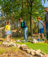 Una familia juega al minigolf al aire libre en un parque vacacional con tiendas glamping y bosque detrás.
