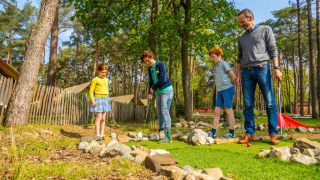 Eine Familie spielt Minigolf im Freien in einem Ferienpark mit Glamping-Zelten und Wald im Hintergrund.
