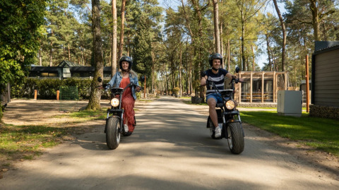 Deux personnes roulent en scooter électrique dans un parc de vacances avec hébergements glamping en arrière-plan.