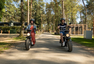 Deux personnes roulent en scooter électrique dans un parc de vacances avec hébergements glamping en arrière-plan.