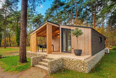 Cabaña moderna para glamping en parque vacacional, rodeada de bosque, con terraza y zona para sentarse.