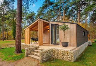Moderne glampinghut in een vakantiepark, omringd door bos, met overdekt terras en buitenzitje.