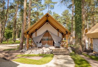 Glampingverblijf op een vakantiepark: luxetent met houten terras en comfortabele zitjes tussen de bomen.