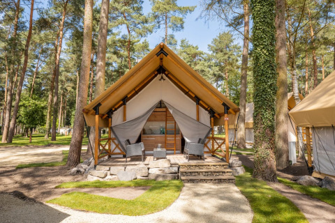 Glampingverblijf op een vakantiepark: luxetent met houten terras en comfortabele zitjes tussen de bomen.