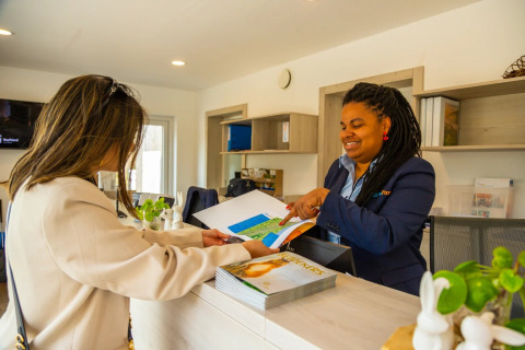 Two women interact at a holiday park reception desk with glamping, reviewing maps and brochures together.