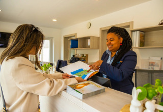 Two women interact at a holiday park reception desk with glamping, reviewing maps and brochures together.