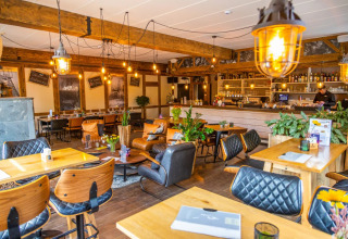 Cozy holiday park lounge with glamping vibe, wooden furniture, plants, pendant lights, and a bar counter.