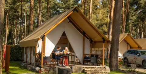 Dos personas disfrutan de bebidas frente a una tienda glamping en un parque vacacional entre pinos.