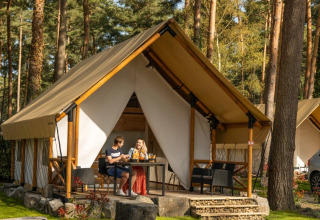 Twee personen genieten van een drankje voor een glampingtent in een vakantiepark omringd door bos.