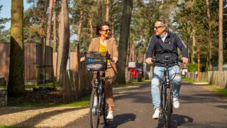 Zwei Menschen fahren Fahrrad in einem Ferienpark mit Bäumen und modernen Glamping-Unterkünften.