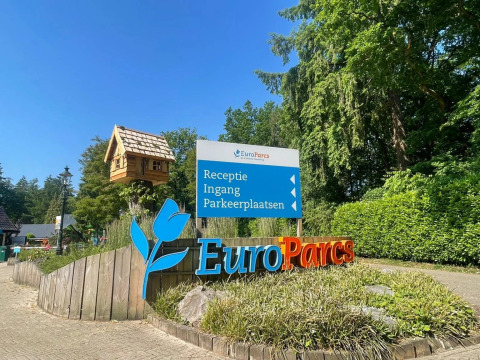 Panneau d'entrée du parc de vacances EuroParcs entouré de verdure, ciel bleu et une grande cabane.
