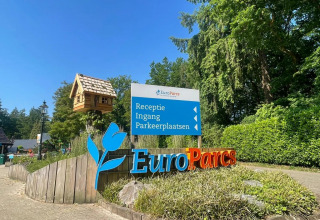 Indgangsskilte ved EuroParcs feriepark med grønne omgivelser og træhytte under blå himmel.