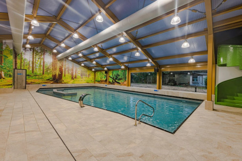 Piscina cubierta en parque glamping, mural de bosque, tobogán verde, ventanas grandes, suelo de baldosas.
